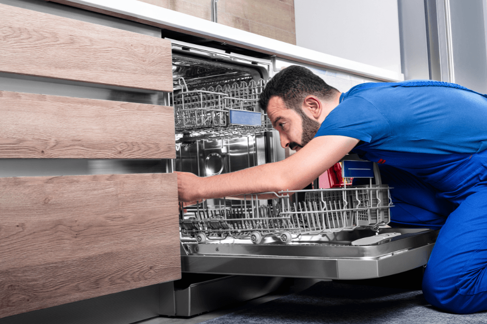 A plumbing technician kneeling at an open built-in dishwasher in a modern kitchen with wood-effect cabinets, reaching in behind the lower rack to check the water supply and drain connections during a dishwasher installation in Dubai
