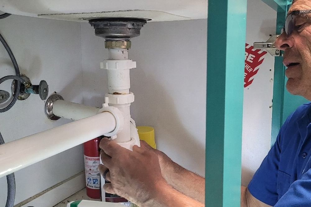 Elderly Quick Fix Dubai technician wearing safety glasses, working underneath a bathroom sink cabinet to reassemble a white PVC P-trap and drain assembly, with a fire extinguisher and yellow bucket visible in the cabinet beside him