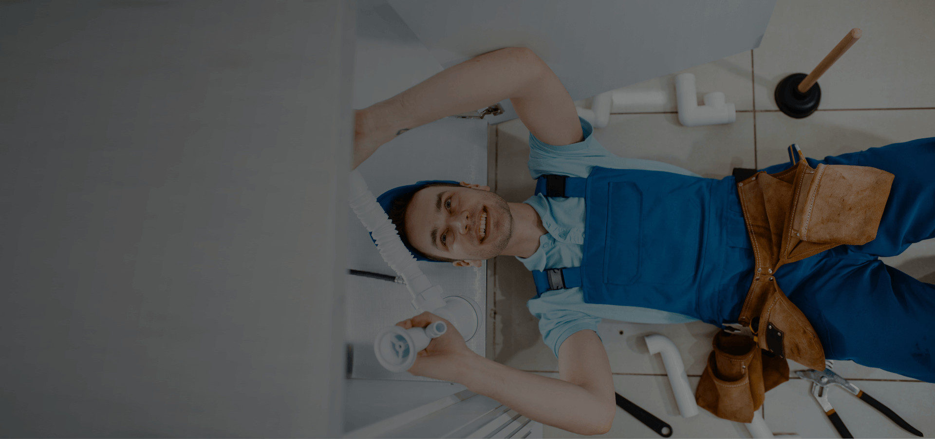 Smiling Quick Fix Dubai plumber wearing a leather tool belt, kneeling in a residential bathroom holding a section of white PVC pipe fitting beside an open sink cabinet, with a toilet plunger and spare pipe fittings visible on the floor