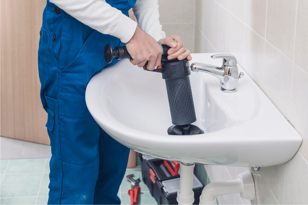 Quick Fix Dubai plumber in blue uniform using a handheld pneumatic drain-clearing tool on a bathroom sink in Dubai, demonstrating manual pressure release for a stubborn clog before escalating to full high-pressure hydro jetting equipment
