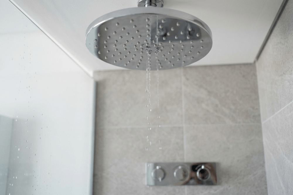 A ceiling-mounted rain shower head in a modern Dubai bathroom giving only a few thin, weak trickles of water from a handful of nozzles while most of the nozzles stay dry, a typical sign of low water pressure in an apartment or villa water supply