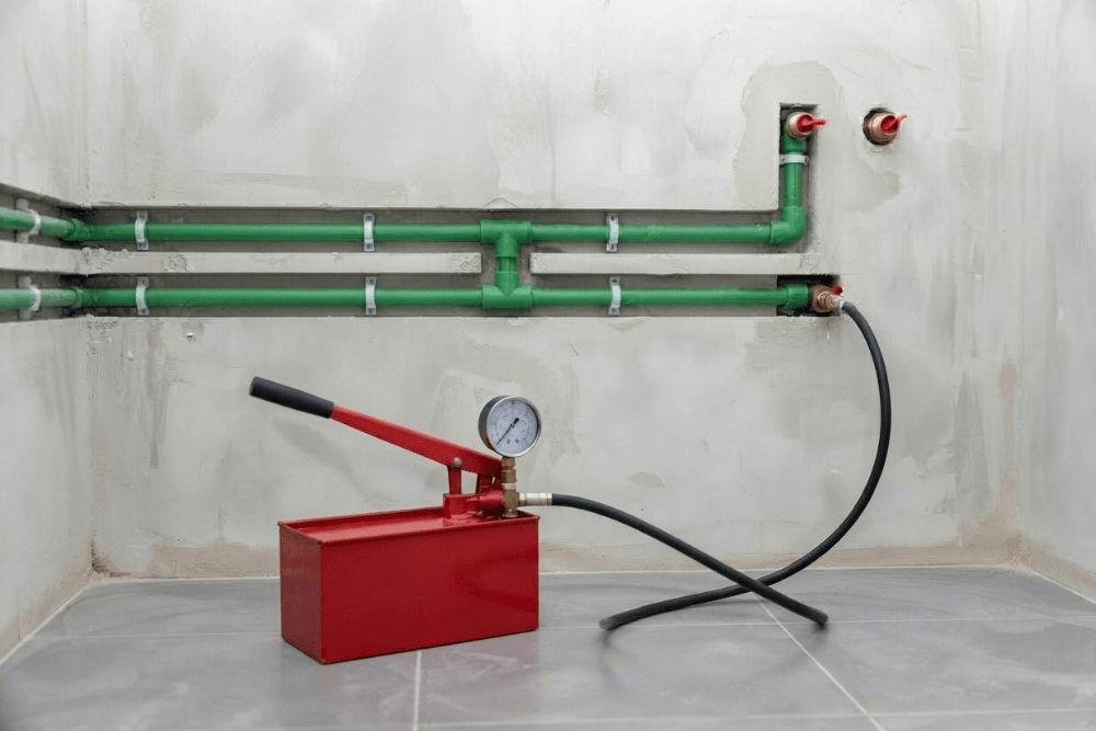 New green PPR hot and cold water pipes fixed with clips in open wall chases during a Dubai bathroom renovation, ending at brass outlets with red-handled valves, with a red hand-operated hydrostatic test pump and pressure gauge connected by a black hose to pressure test the new pipework before the walls are closed