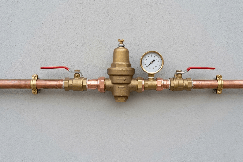 A brass pressure reducing valve with an adjustment screw on its bell-shaped top and a pressure gauge on its outlet side, installed on a copper main water supply pipe clipped to a villa wall in Dubai, with a red-handled ball valve on each side so the PRV can be isolated for adjustment or replacement