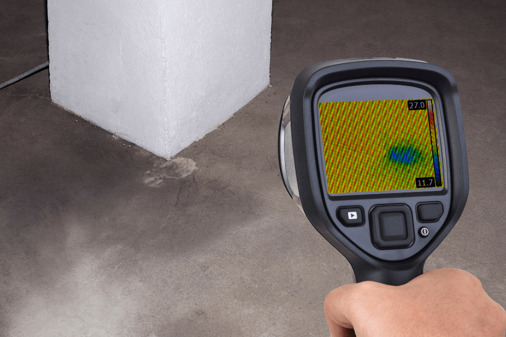 Handheld thermal infrared imaging camera displaying a colour heat-map on its screen, aimed at a damp patch on a concrete slab floor beside a white structural column, used to visually detect a temperature anomaly consistent with a hidden slab leak before any demolition begins